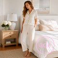 A woman stands barefoot in a bedroom wearing a short-sleeve button-front pajama top and matching plaid pajama pants. The set is light cream with a subtle pale yellow grid pattern and pale yellow piping.