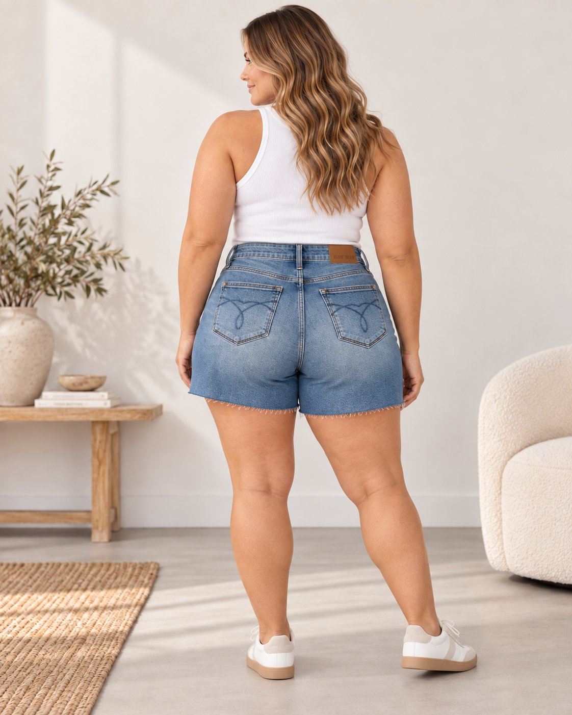 A woman wearing light-wash denim shorts and a white tank top, shown from the back in a bright, modern indoor room with a chair, side table, and plant.