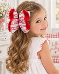 A young girl with long wavy light brown hair wearing a large red and white patterned hair bow, photographed in a bright indoor room setting.
