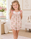 A young girl stands in a bright nursery wearing a sleeveless romper with a pink and green floral pattern, a white collar personalized with the name 'Ellie', and ruffled shoulder ties. The room has a white wall, a framed sign, a bunny plush, and a side table with flowers.