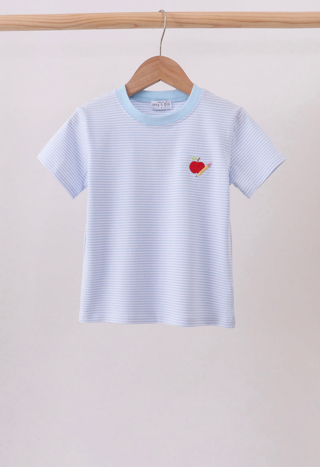 A light blue and white striped short-sleeve t-shirt with a small red embroidered apple on the chest, hanging on a wooden hanger from a wooden clothes rack against a plain off-white wall.
